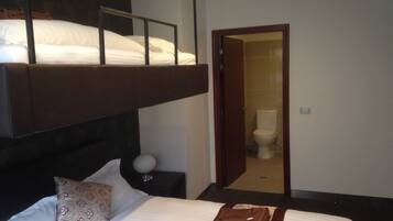 A hotel room with a bunk bed, a single bed, a bedside table with a lamp, and a bathroom visible through an open door.