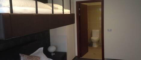 A hotel room with a bunk bed, a single bed, a bedside table with a lamp, and a bathroom visible through an open door.