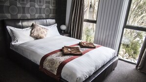 A neatly made bed with a patterned throw, a bedside table with a lamp, and a view of trees through large windows. - Duck Inn Mt Buller (Mount Buller)