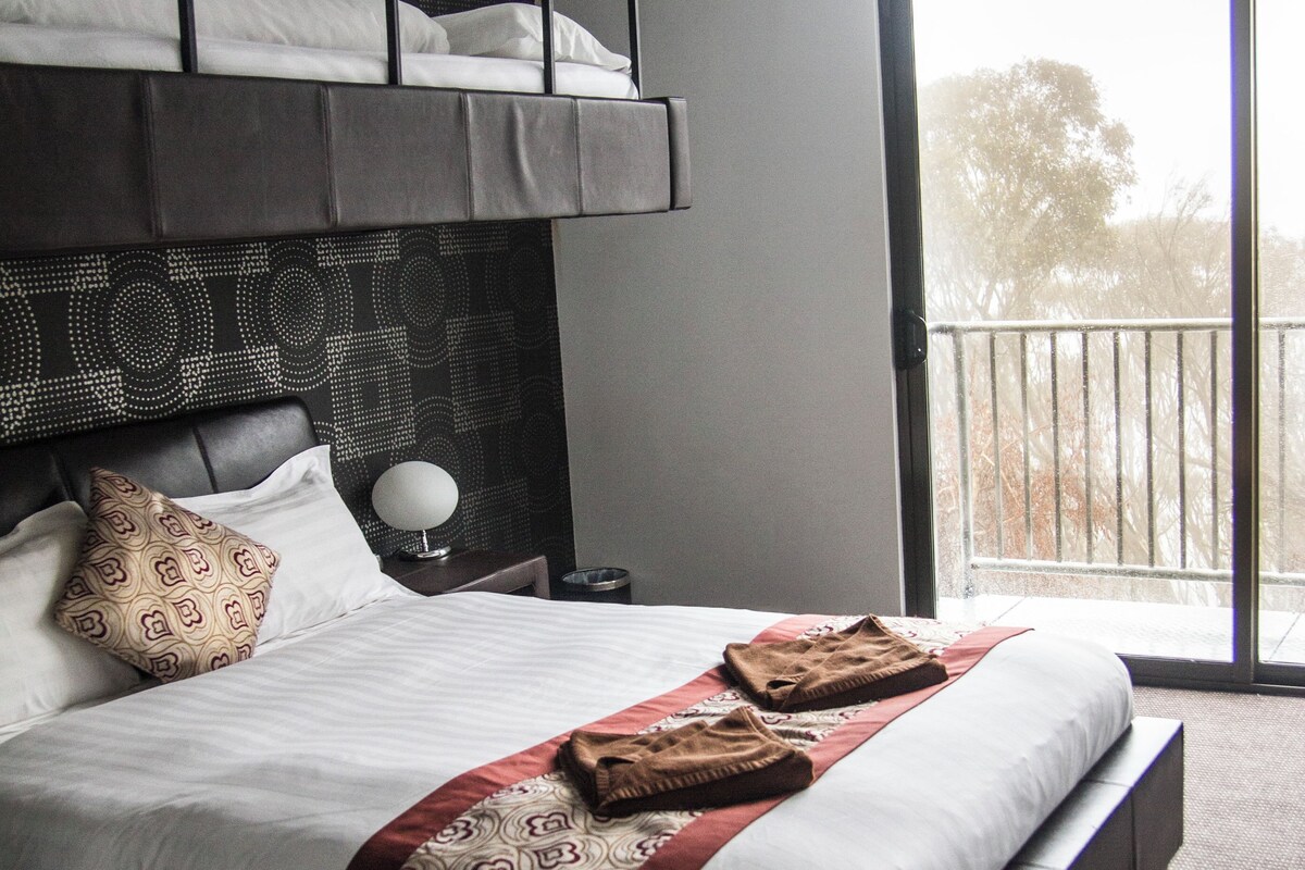 A modern hotel room with a large bed, a headboard with a geometric pattern, a bedside table with a lamp, and a balcony with a railing.