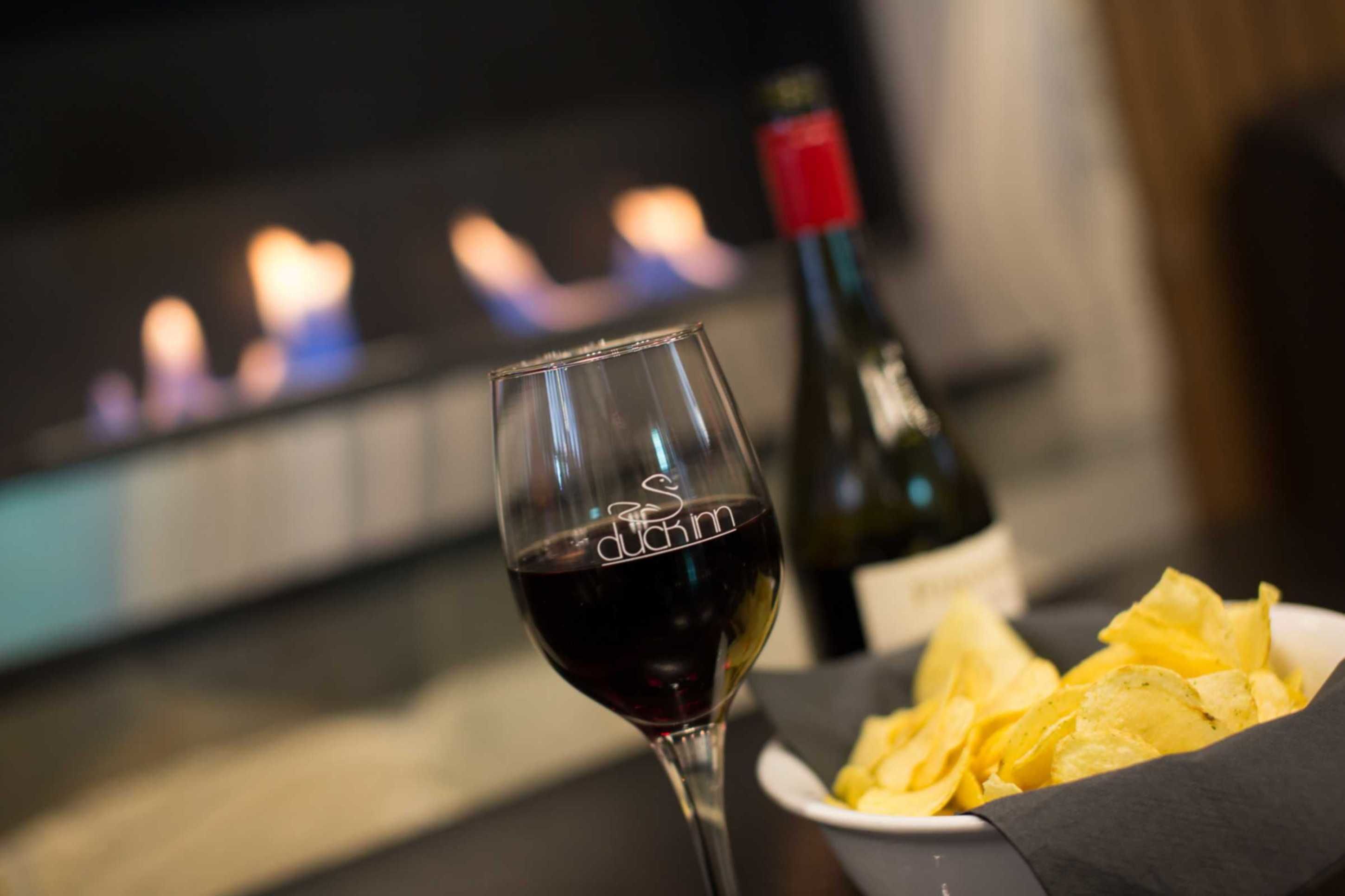 A glass of red wine, a bottle of wine, and a plate of chips on a table with a fireplace in the background.