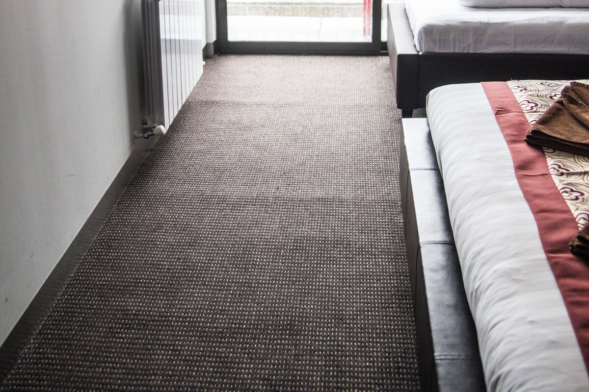 A hotel room with a bed, a TV, a radiator, a sliding glass door to a balcony, and a view of trees outside.