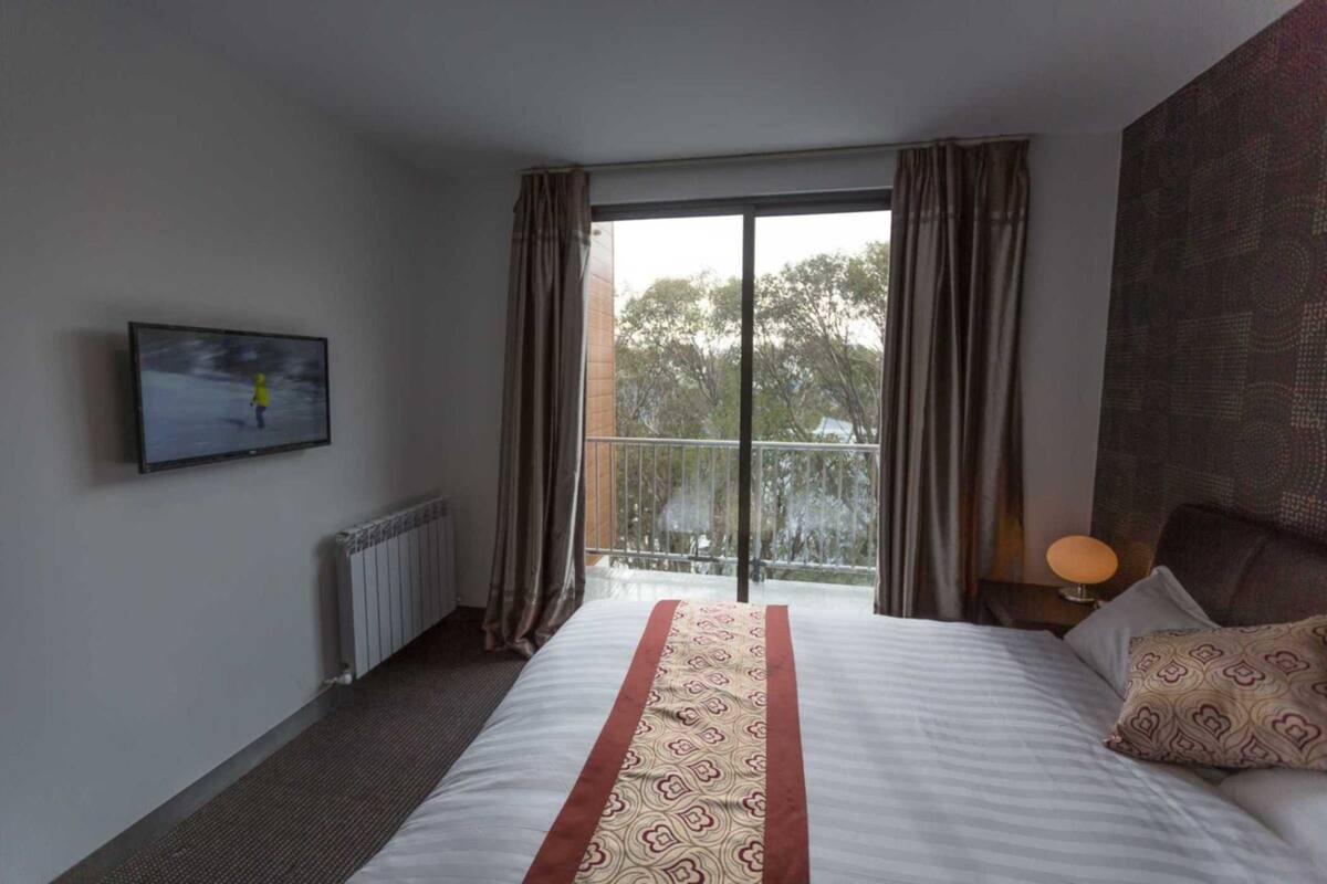 A hotel room with a large bed, a television on the wall, a balcony with curtains, and a view of trees.