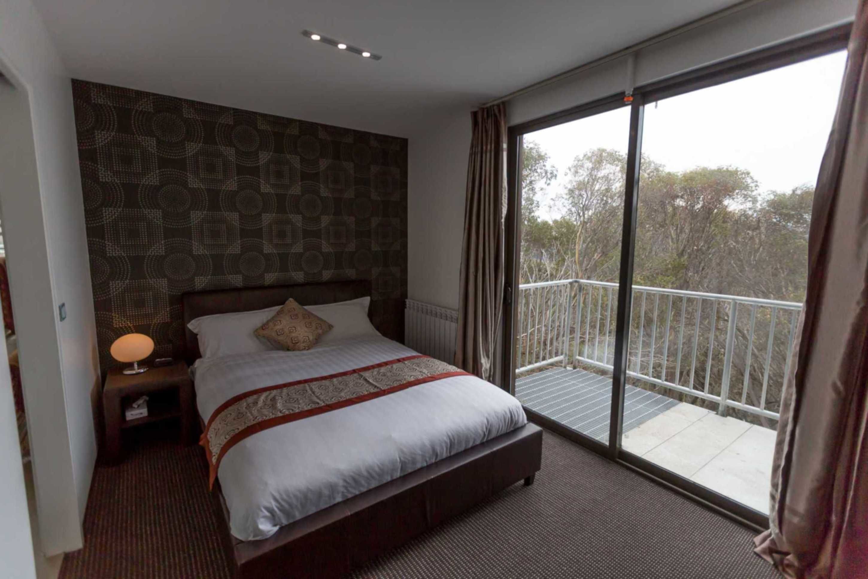 A hotel room with a large bed, a nightstand, a lamp, and a view of the outdoors through a sliding glass door.
