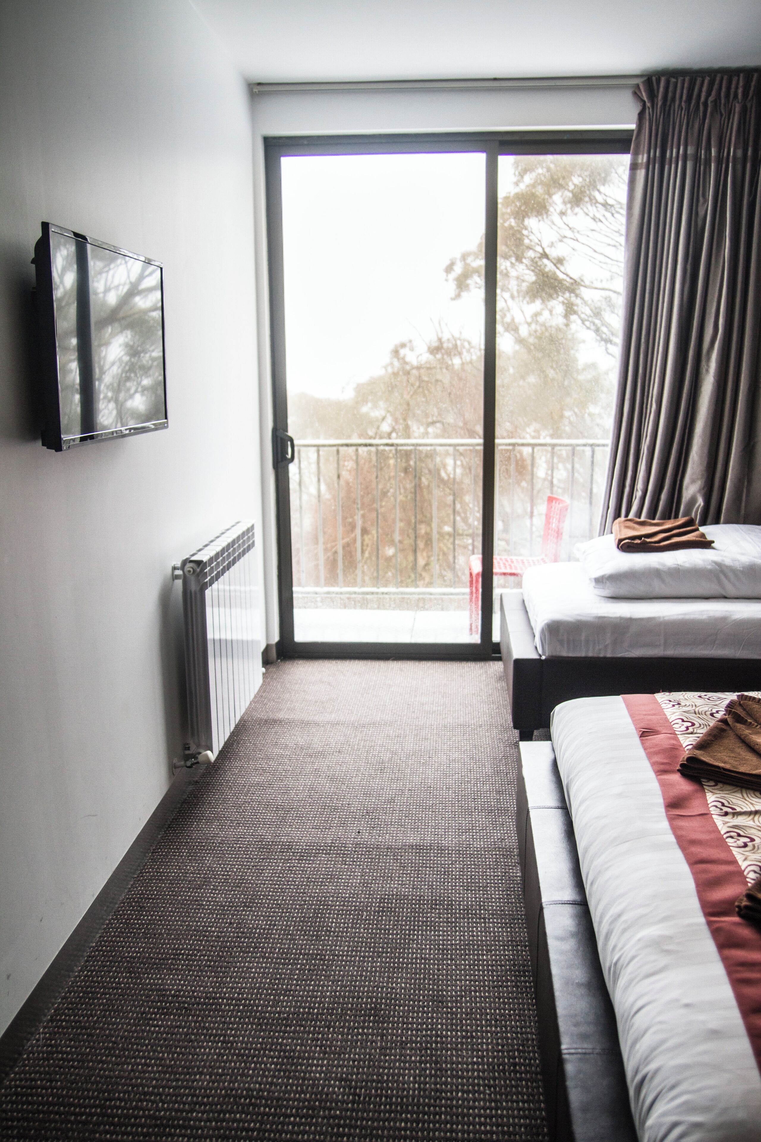 A hotel room with a bed, a TV, a radiator, a sliding glass door to a balcony, and a view of trees outside.