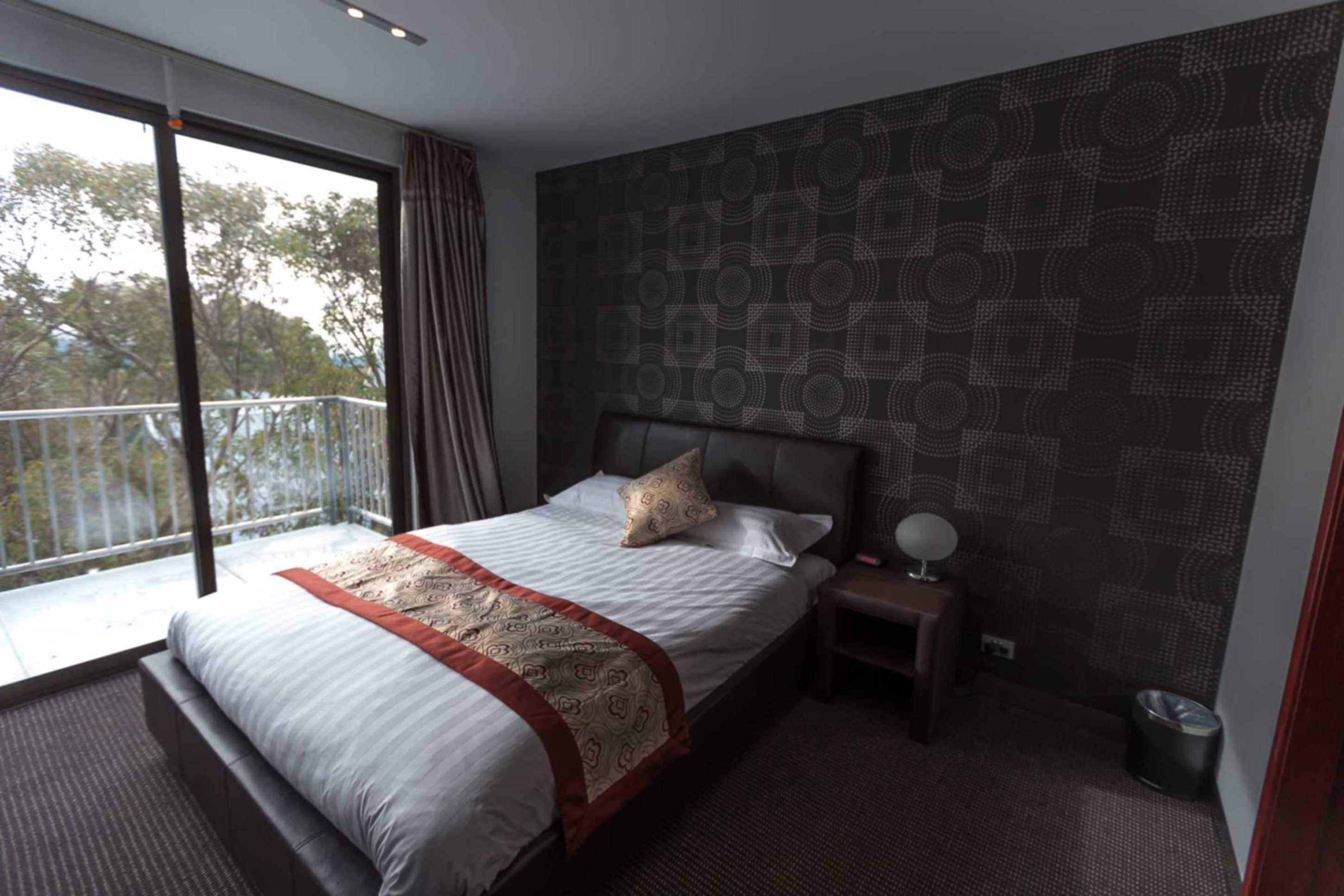 A modern hotel room with a large bed, a bedside table with a lamp, a view of the outdoors through a balcony, and a patterned wall.