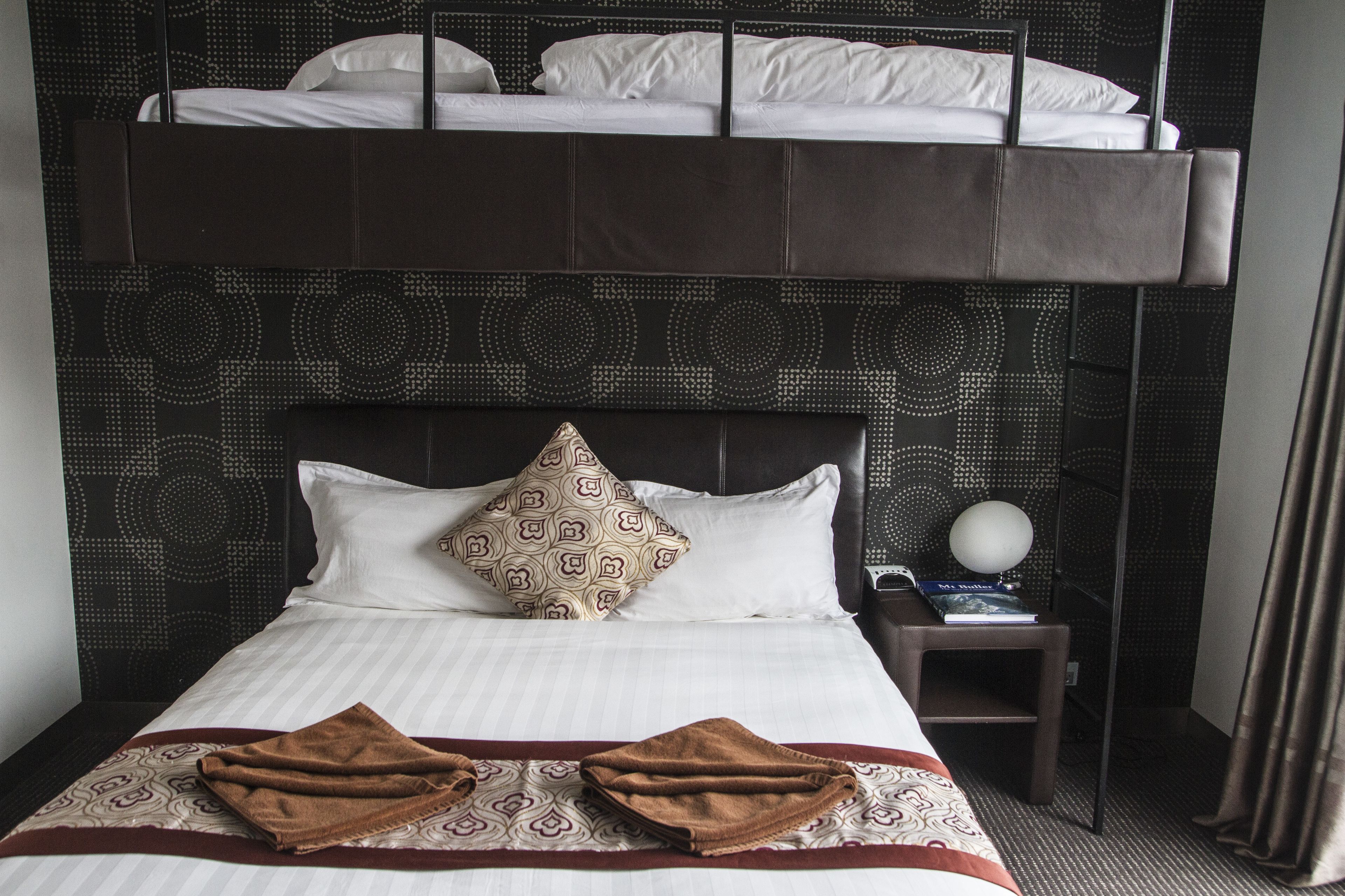 A hotel room with a bunk bed, a patterned headboard, a bedside table with a lamp, and a neatly made bed with towels.