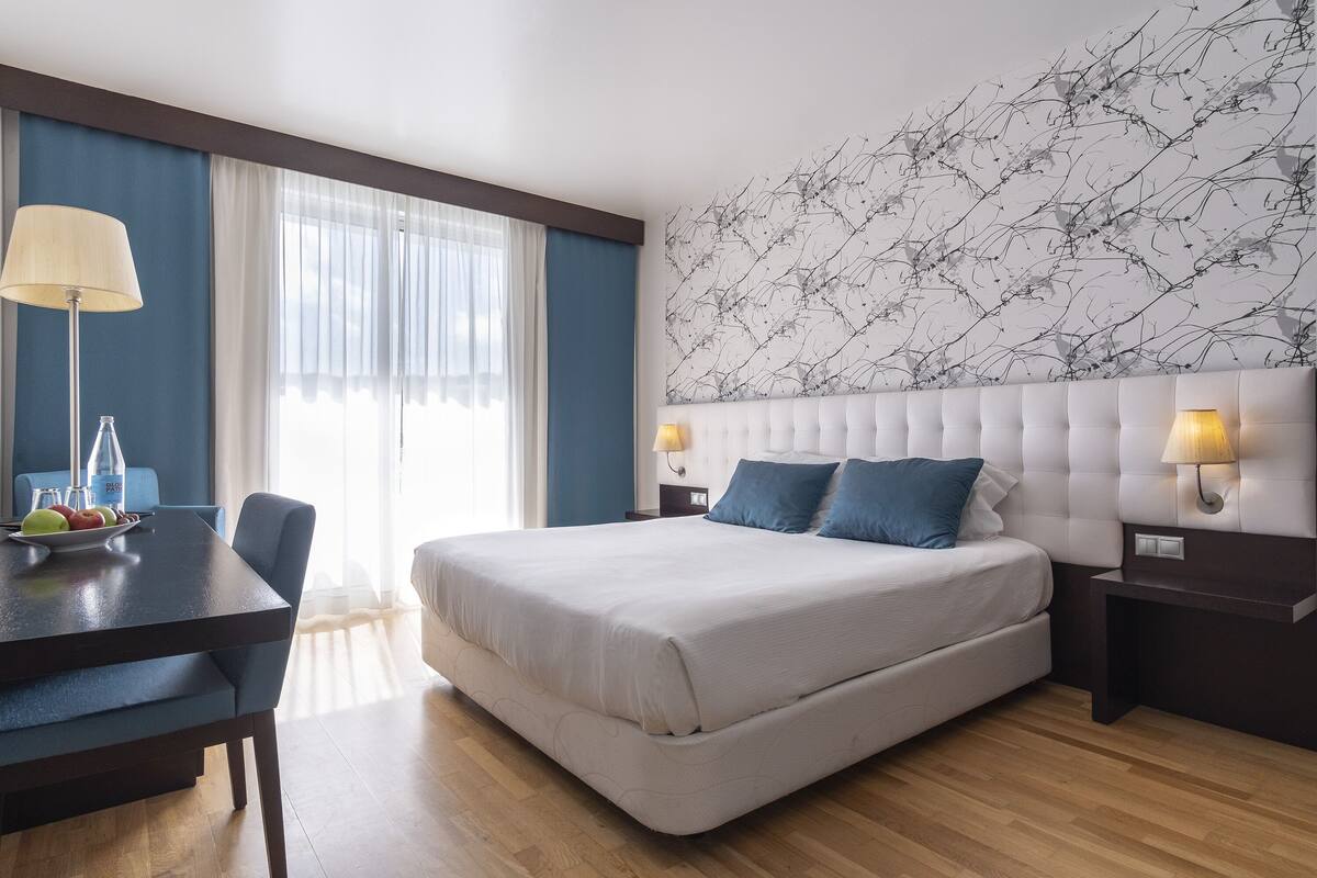 A hotel room with a large bed, a desk with a lamp, a chair, and a view of the outdoors through the window.