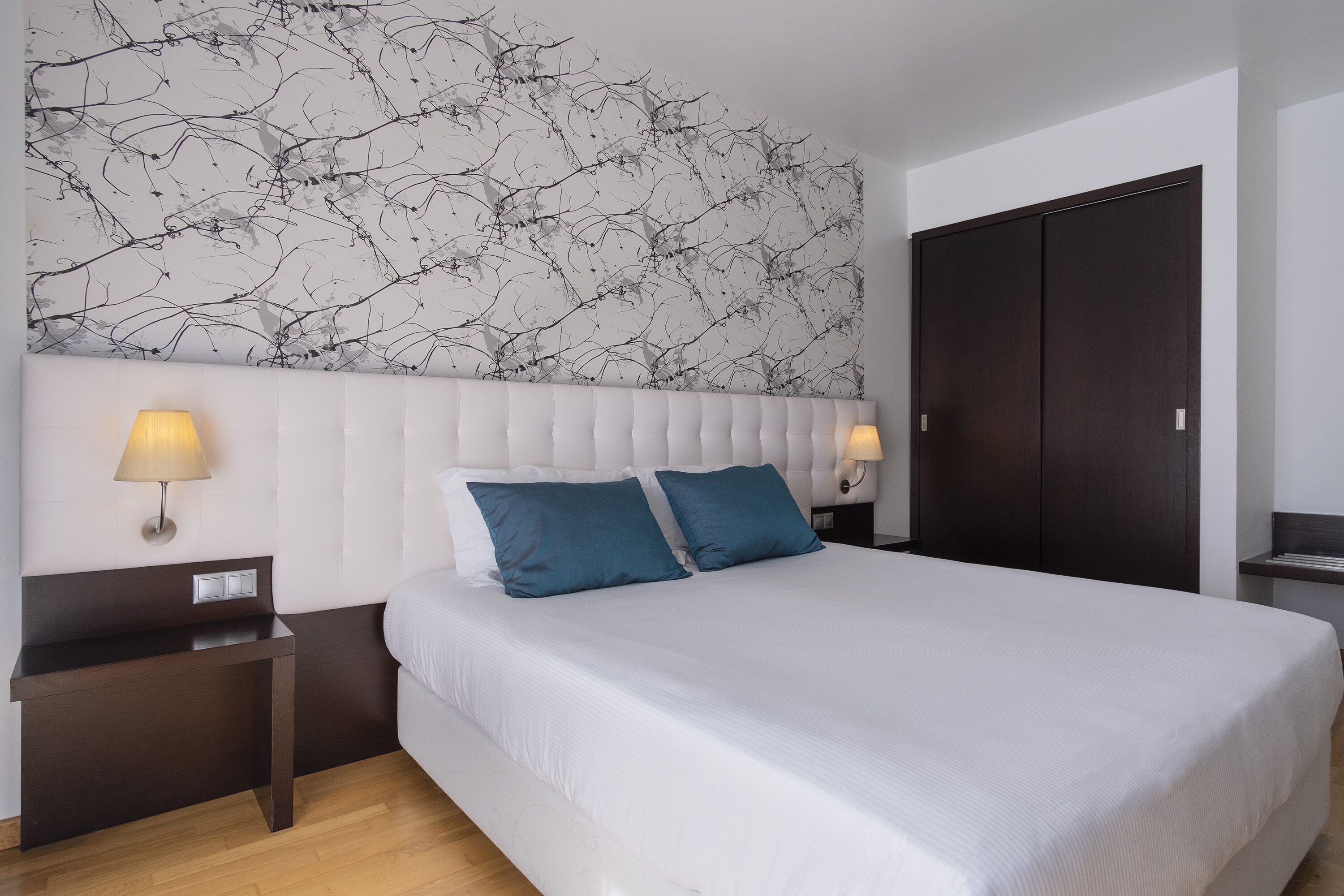 Quarto com uma cama grande, duas luminárias de cabeceira, uma mesa de cabeceira de madeira escura e um guarda-roupa escuro.