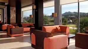 A modern hotel lobby with orange sofas, large windows offering a view of a golf course and mountains, and floor-to-ceiling glass doors. - Hotel Vale do Navio (Ponta Delgada)