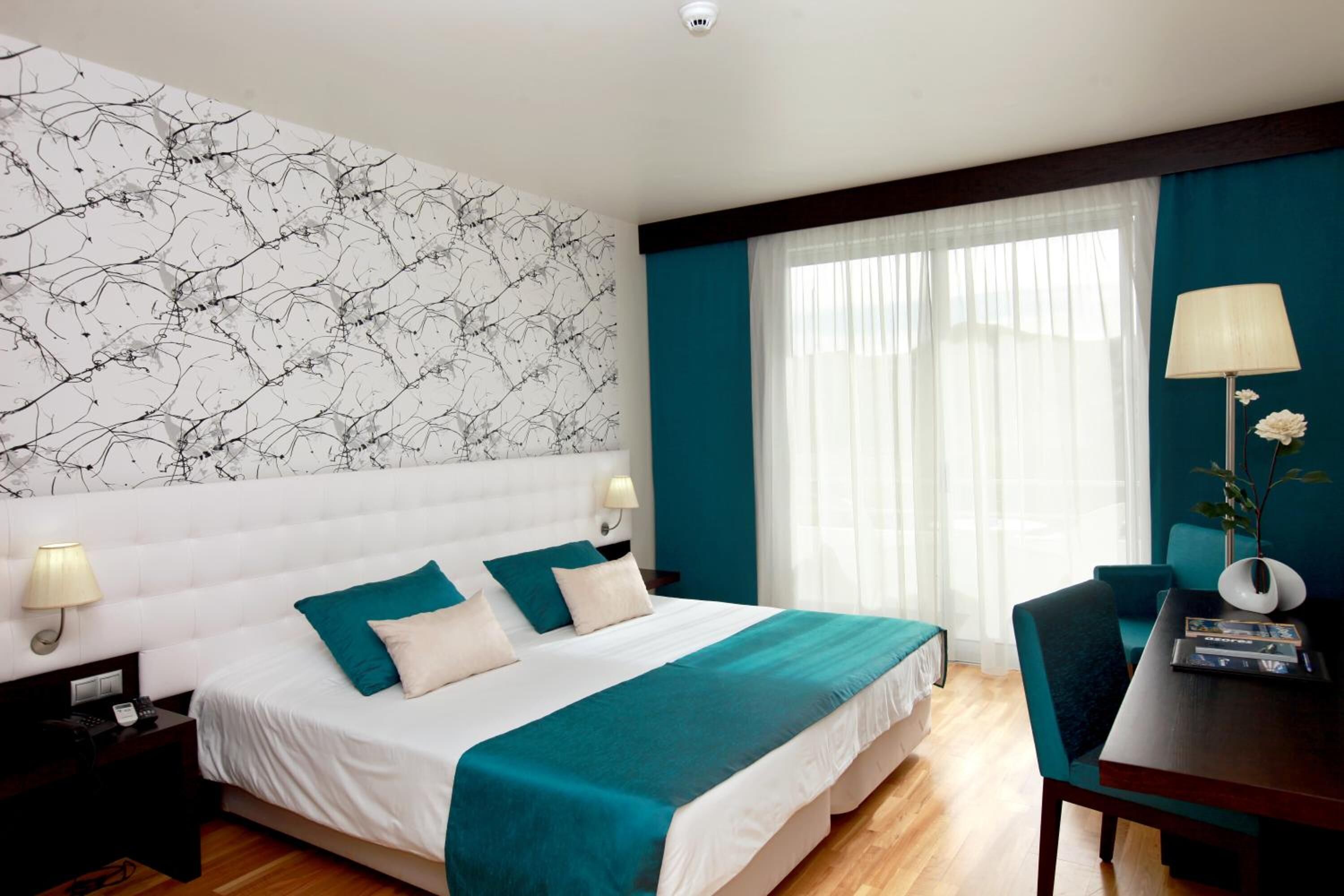 Une chambre d’hôtel avec un grand lit, un bureau avec une chaise, une table de chevet et un mur décoré d’un motif représentant des branches.