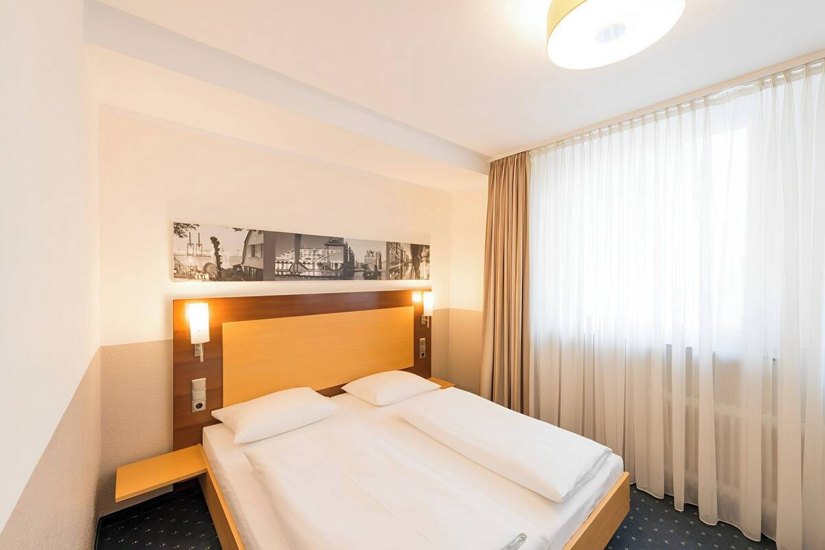 A hotel room with a neatly made bed, a headboard with a cityscape artwork, a bedside table, and a window with sheer curtains.