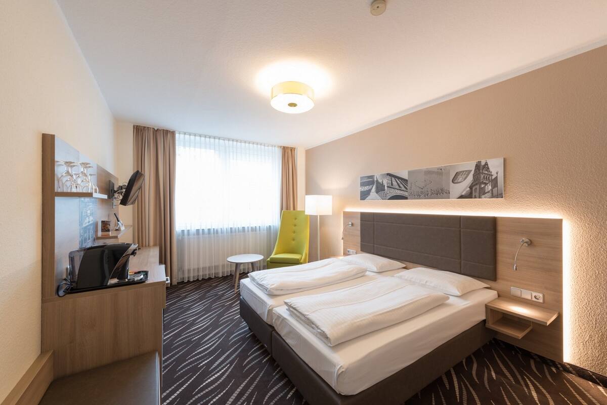 A hotel room with a bed, a desk, a chair, a lamp, and artwork on the wall.