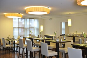 A modern dining area with multiple tables and chairs, large windows, and pendant lighting.