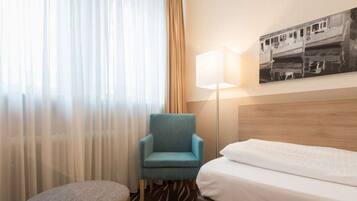 A hotel room with a bed, a blue armchair, a small ottoman, a floor lamp, and a painting on the wall.