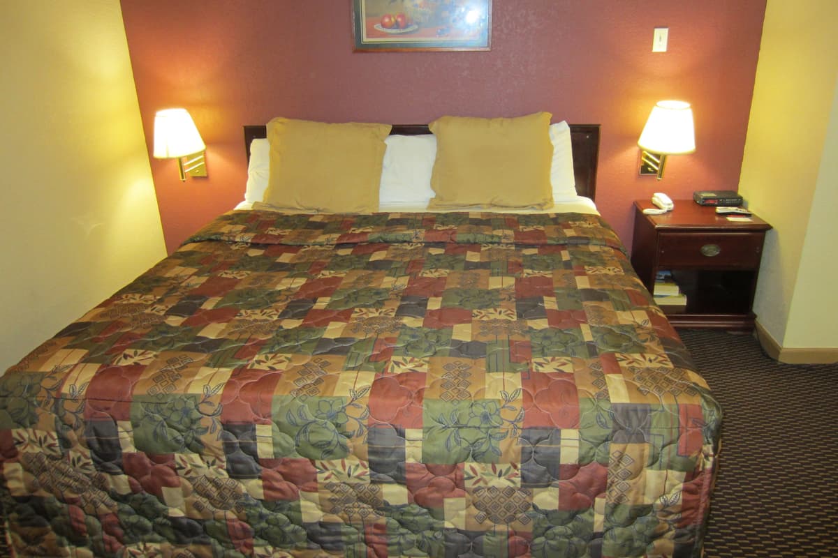 A hotel room with a large bed, two bedside lamps, a nightstand with a phone and remote controls, and a painting on the wall.