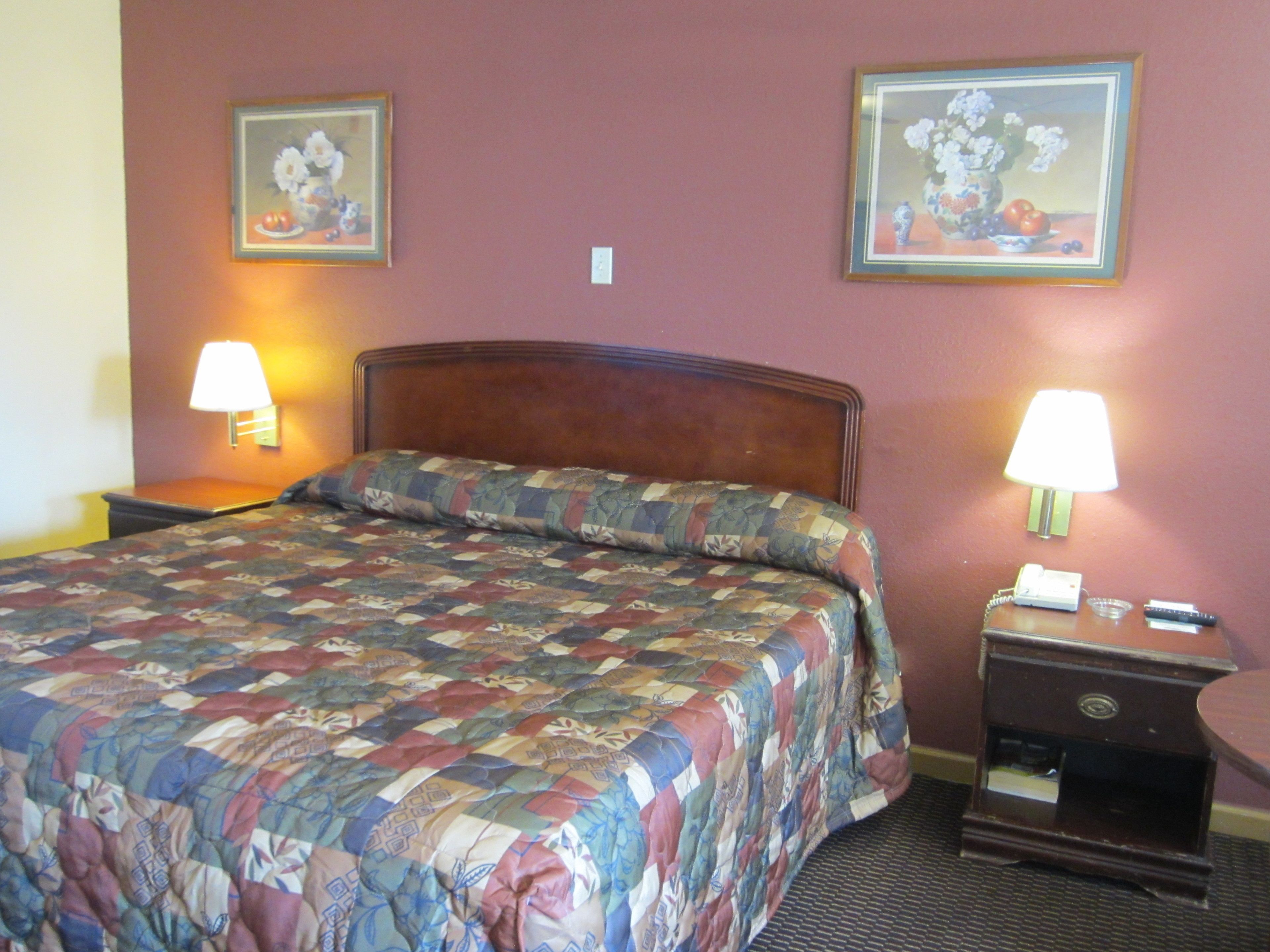 Una habitación de hotel con una cama, dos lámparas de noche, una mesita de noche con un teléfono y dos cuadros enmarcados con motivos florales en la pared.
