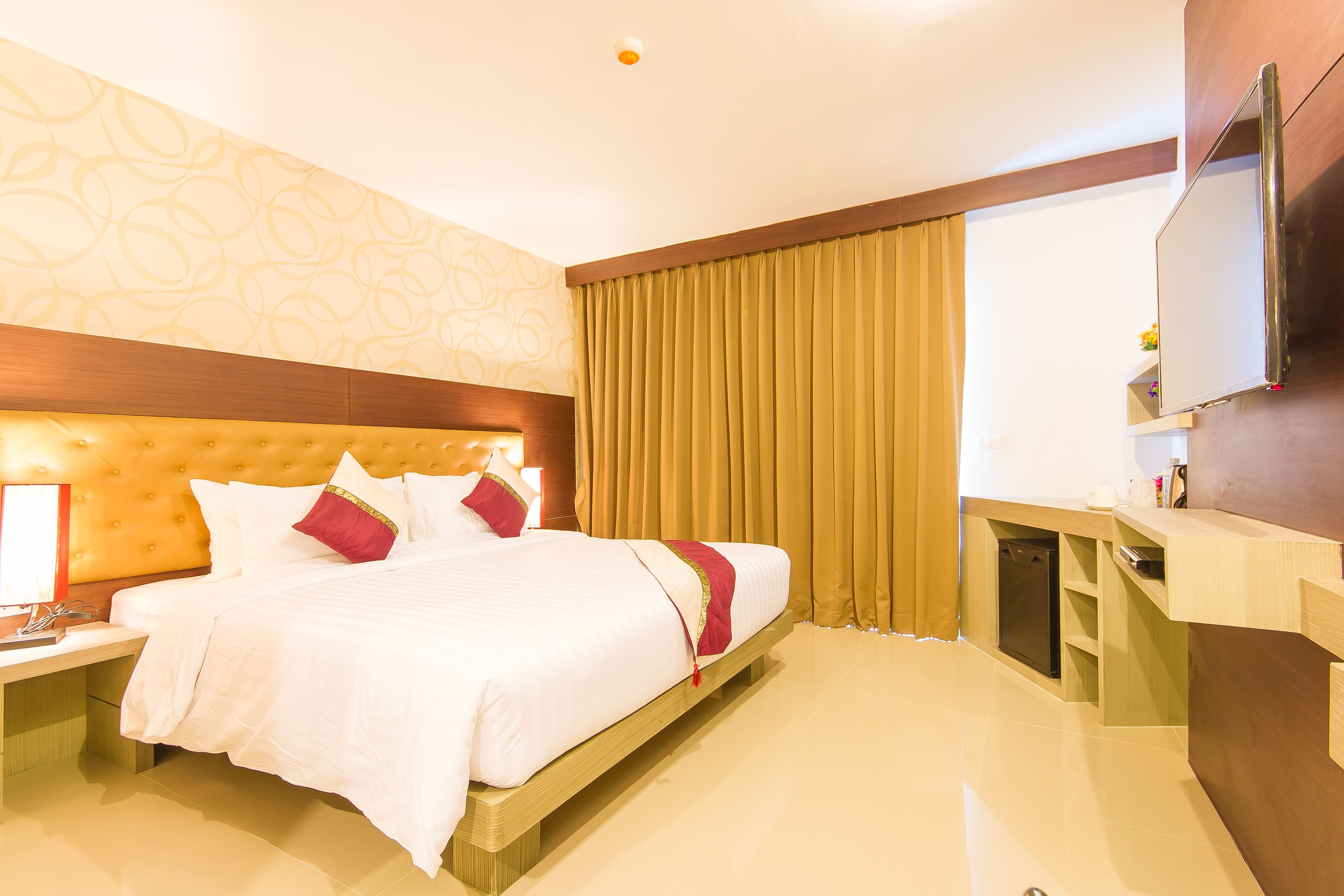 A hotel room with a large bed, a wooden headboard, a bedside table with a lamp, a built-in TV, and a small seating area with a chair and table.