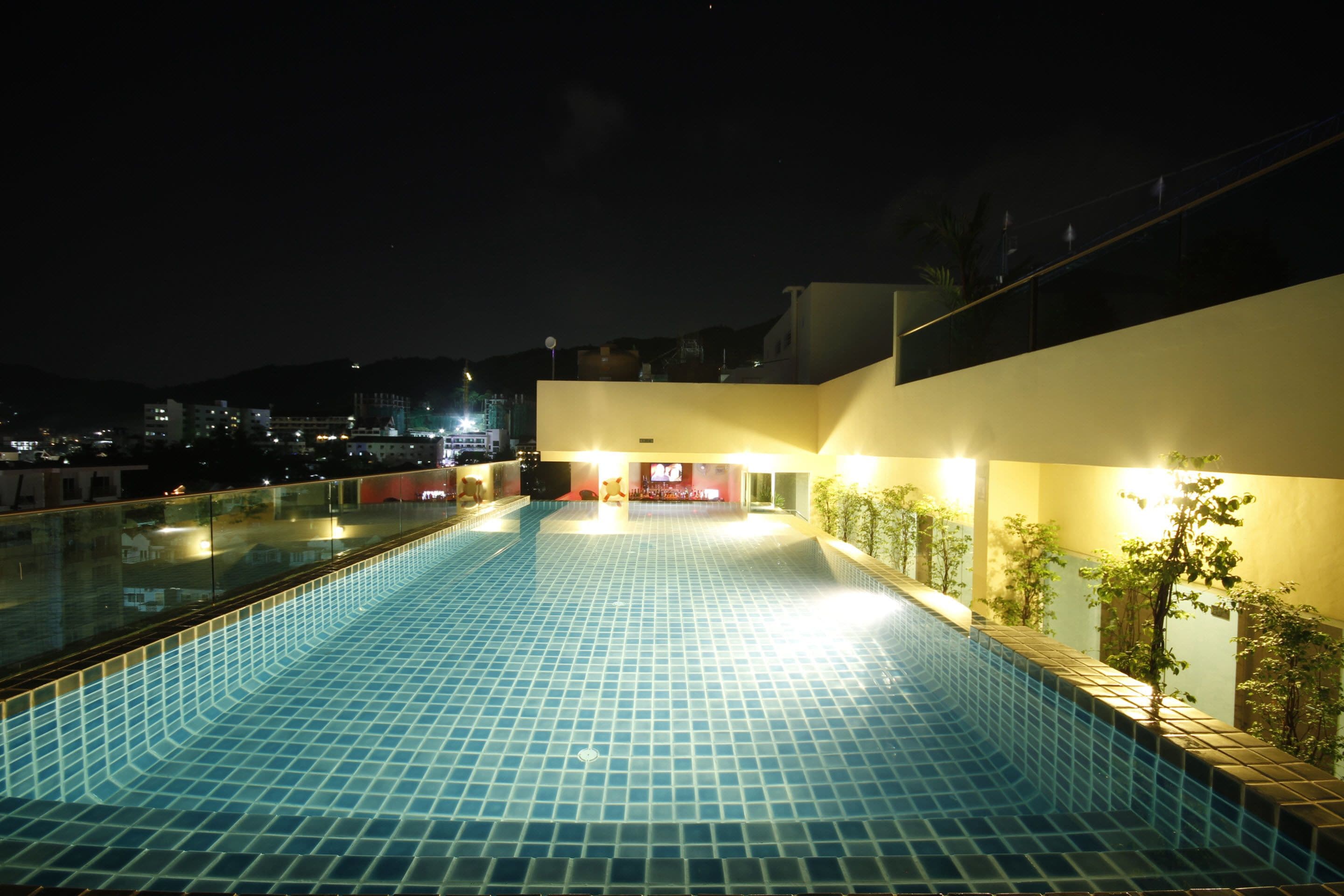 A swimming pool with tiled flooring and a glass railing, surrounded by walls with small plants and a building in the background.