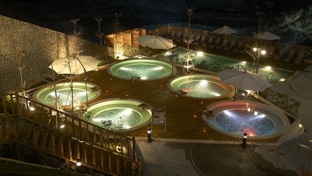 Vista nocturna de un complejo turístico con múltiples jacuzzis y zonas de descanso, rodeado por una terraza de madera y muros de piedra.