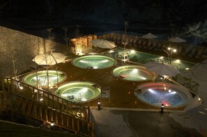 A nighttime view of a resort with multiple hot tubs and lounge areas, surrounded by a wooden deck and stone walls. - Uni Resort KU-KUAN (Taichung)