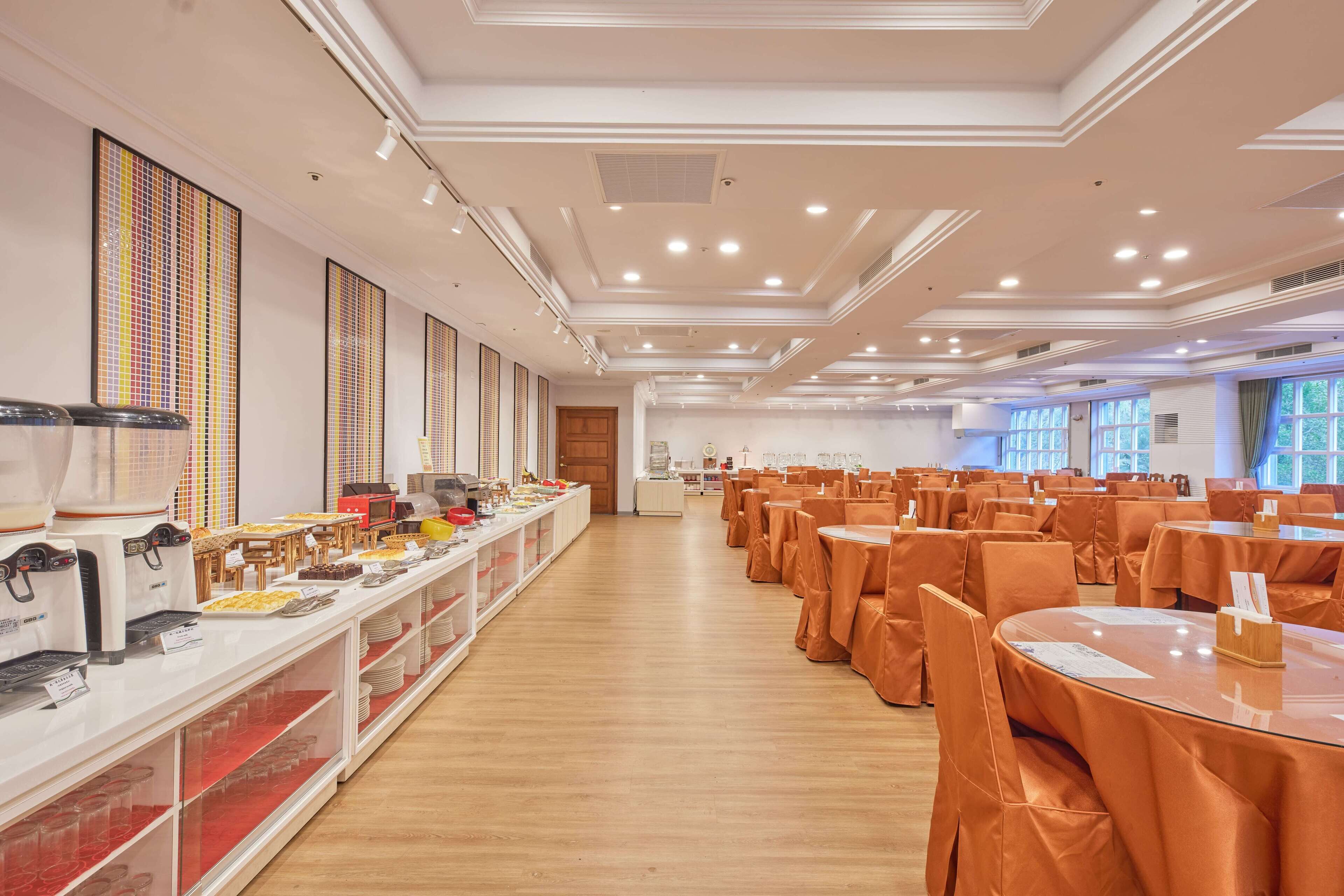 A spacious banquet hall with multiple tables and chairs, a buffet area with various food items, and a modern ceiling design.