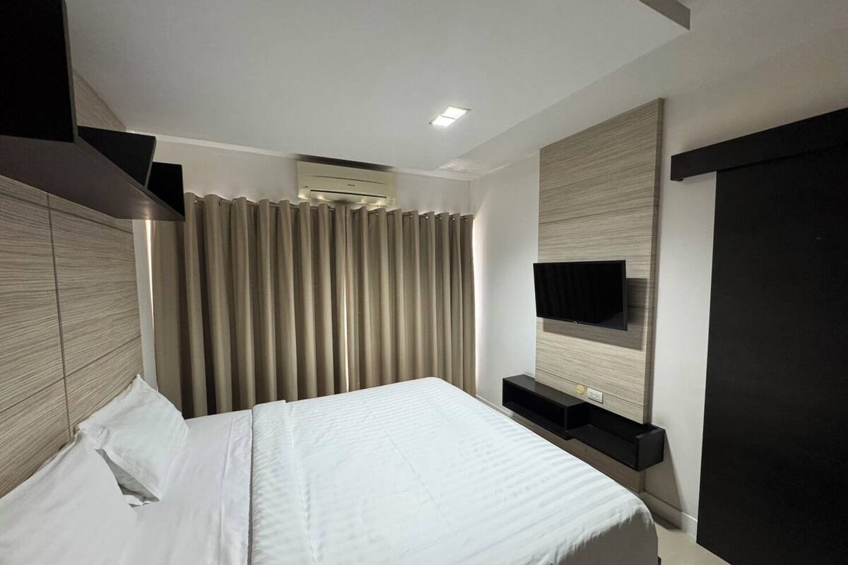 A modern bedroom with a large bed, a flat-screen TV mounted on the wall, and a built-in air conditioning unit.