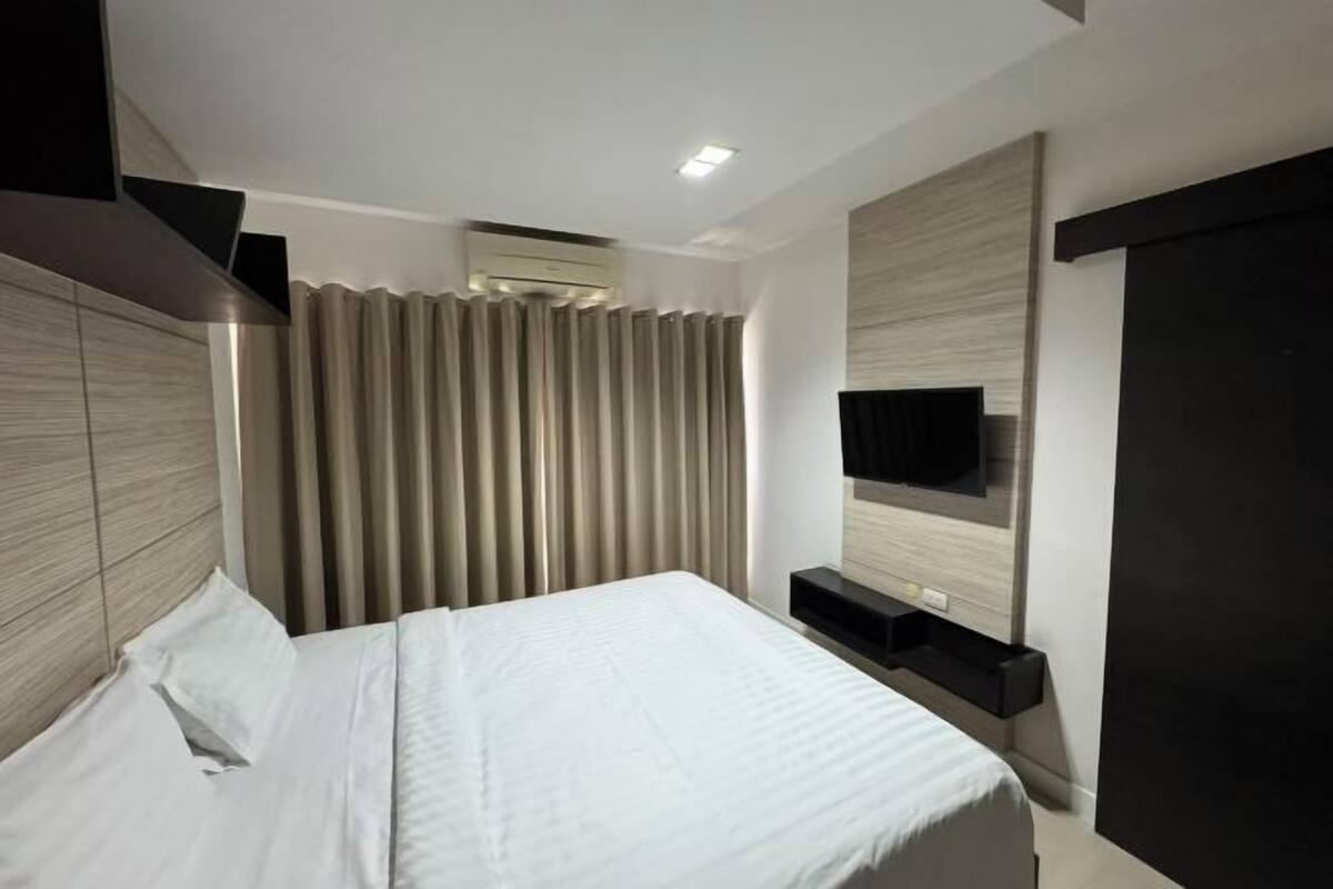 A modern hotel room with a bed, a flat-screen TV mounted on the wall, and a built-in air conditioning unit.