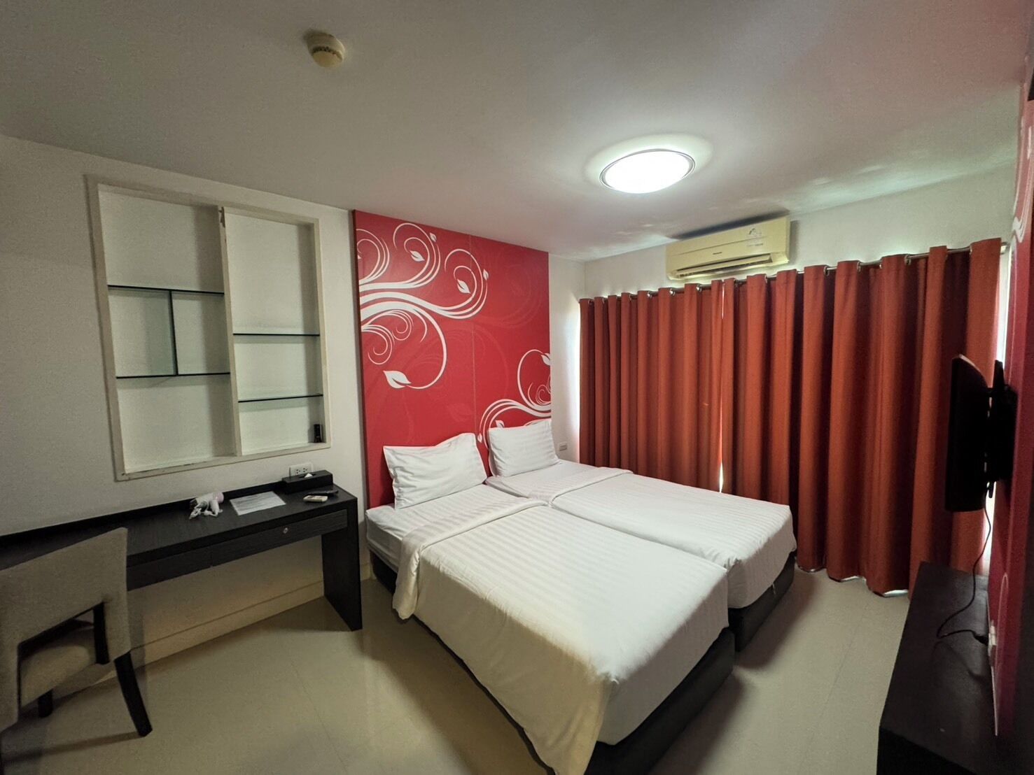 A hotel room with a bed, a desk, a mirror, and red curtains.