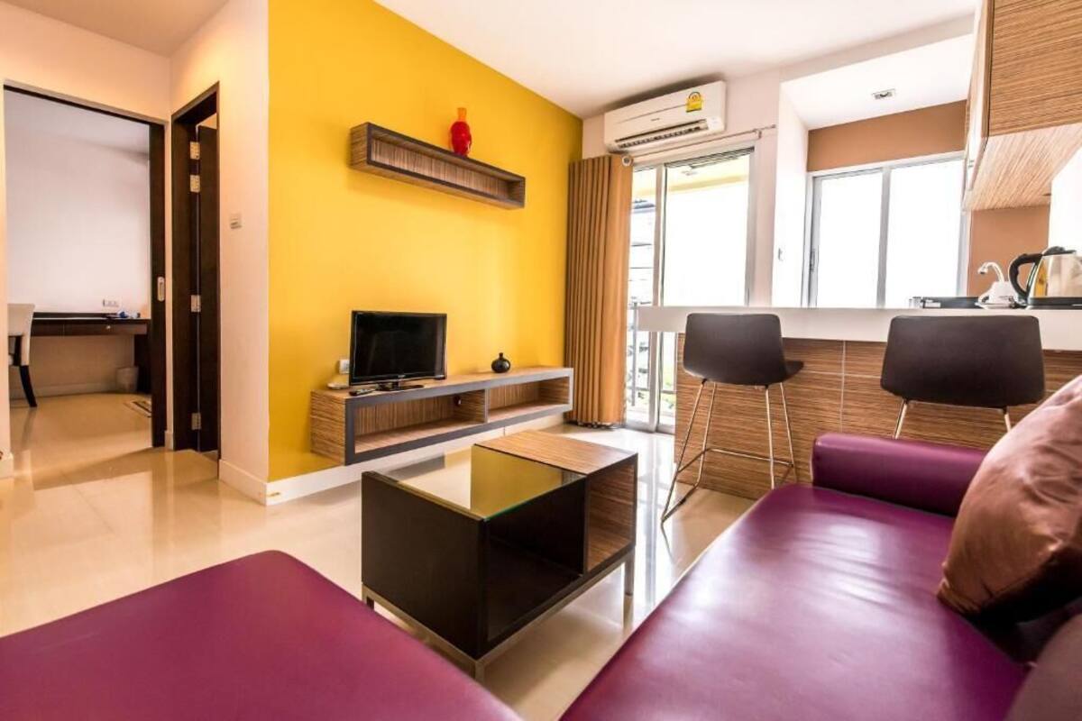 A modern living room with a yellow accent wall, a flat-screen TV, a glass coffee table, and a purple sofa.