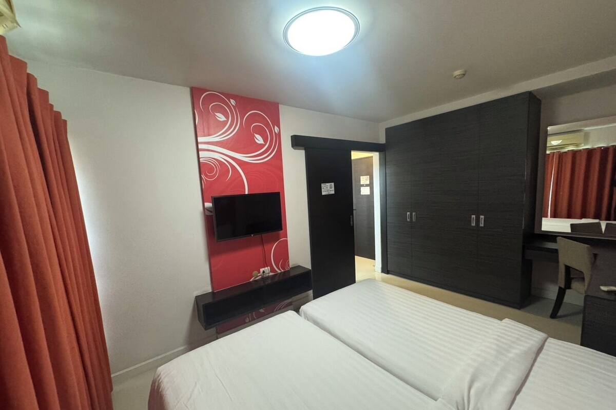 A hotel room with two beds, a TV mounted on a red wall, a wardrobe, and a desk with a mirror.