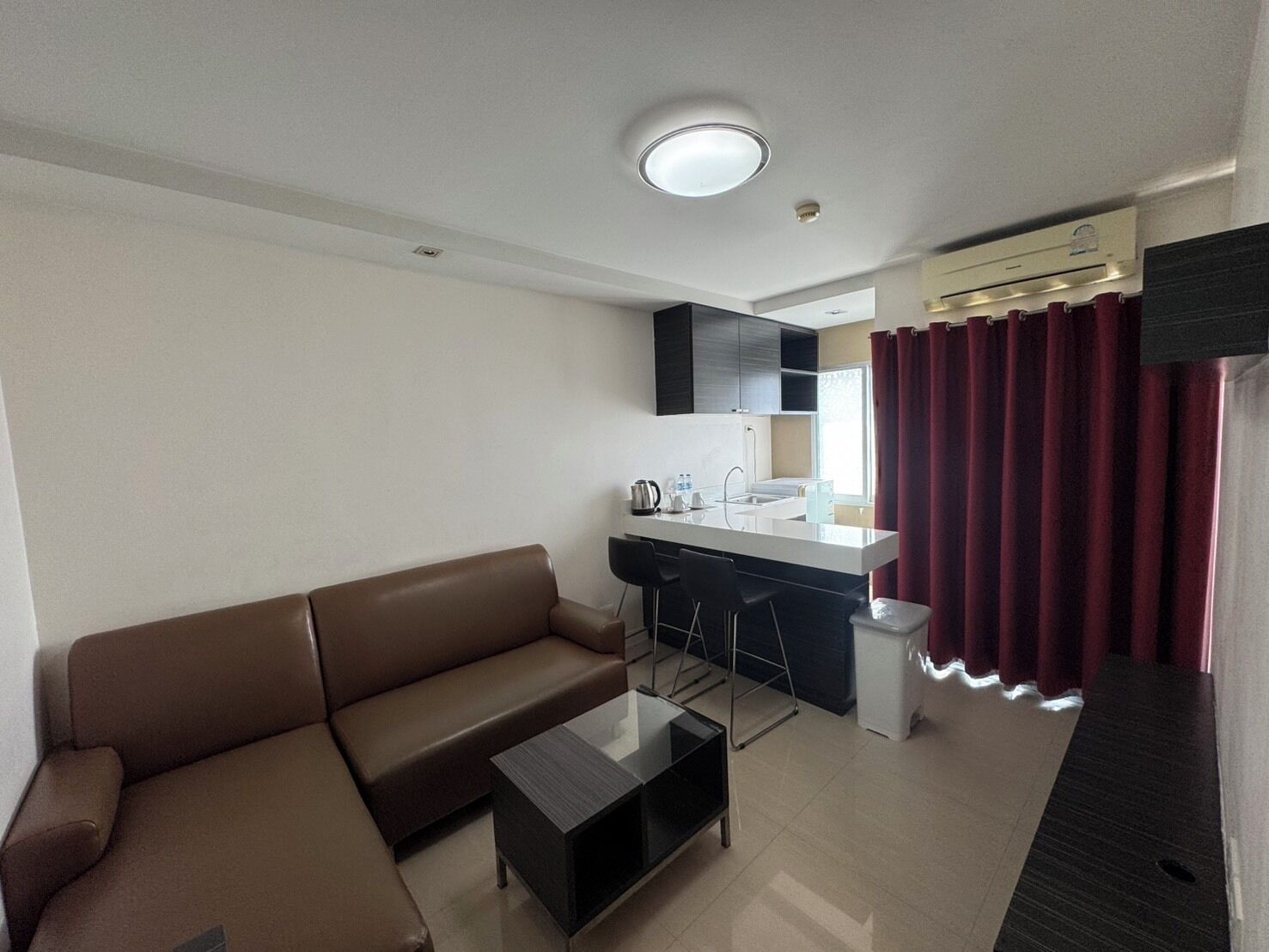 A compact living space with a sofa, a small coffee table, a kitchenette with a bar area, and a window with curtains.