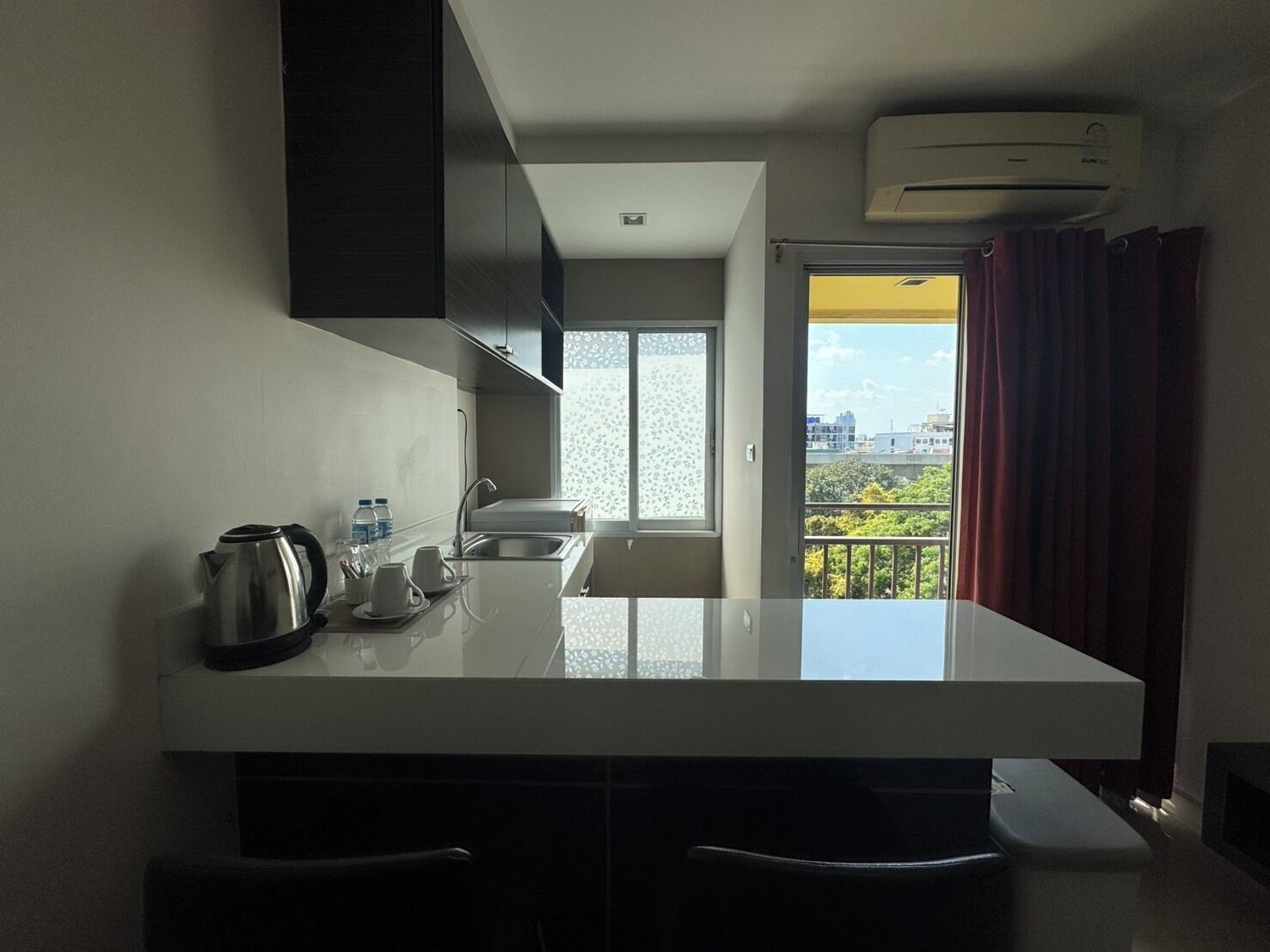 Cocina moderna con encimera blanca, hervidor de acero inoxidable y lavamanos. Se ve una ventana con vistas a edificios y zonas verdes.