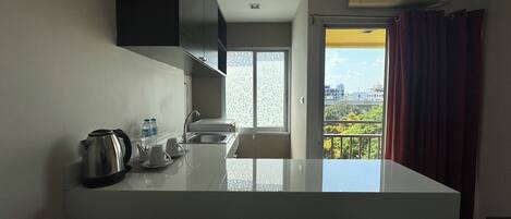 Cocina moderna con encimera blanca, hervidor de acero inoxidable y lavamanos. Se ve una ventana con vistas a edificios y zonas verdes.
