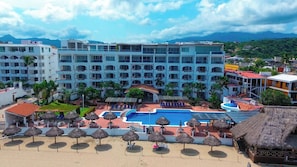 Un resort con piscina, zona de playa con sombrillas y un edificio grande con múltiples balcones.