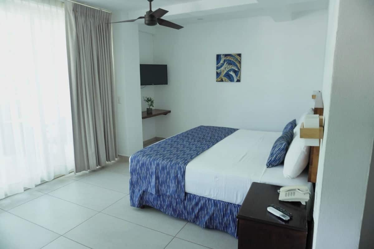 A bedroom with a bed, a ceiling fan, a TV, a bedside table with a phone, and a window with curtains.