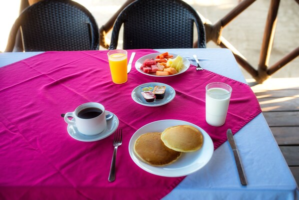 Una mesa dispuesta para el desayuno con tortitas, fruta y bebidas, con un mantel rosa brillante.