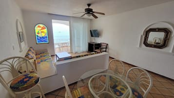 Um quarto aconchegante com um assento junto à janela, paredes brancas, um ventilador de teto, uma TV e um espelho.
