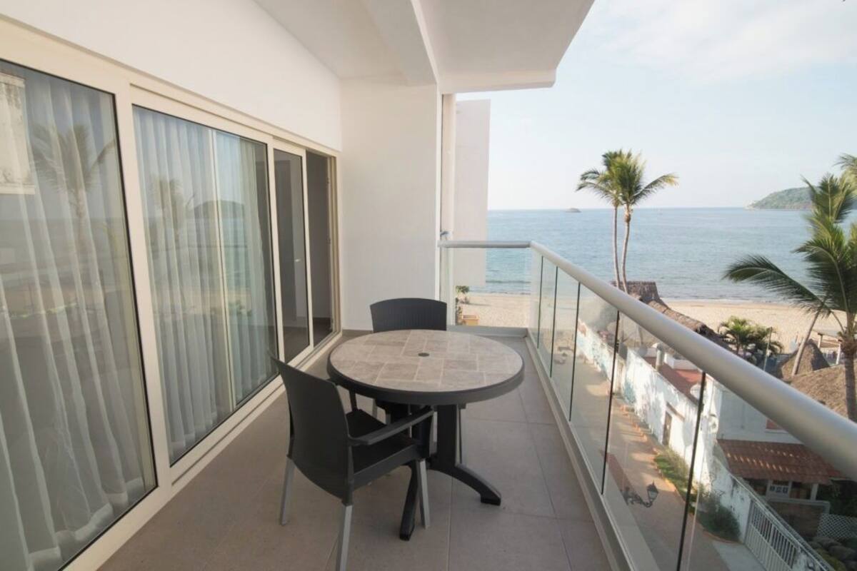 A balcony with a table and chairs, overlooking a beach with palm trees and a view of the sea.