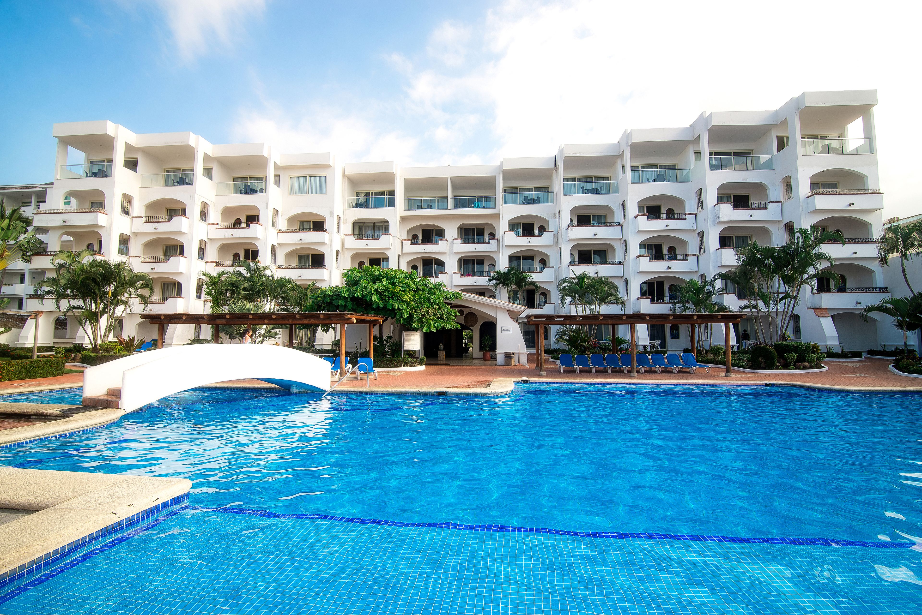 A hotel with a swimming pool, multiple balconies, and a covered area with chairs.
