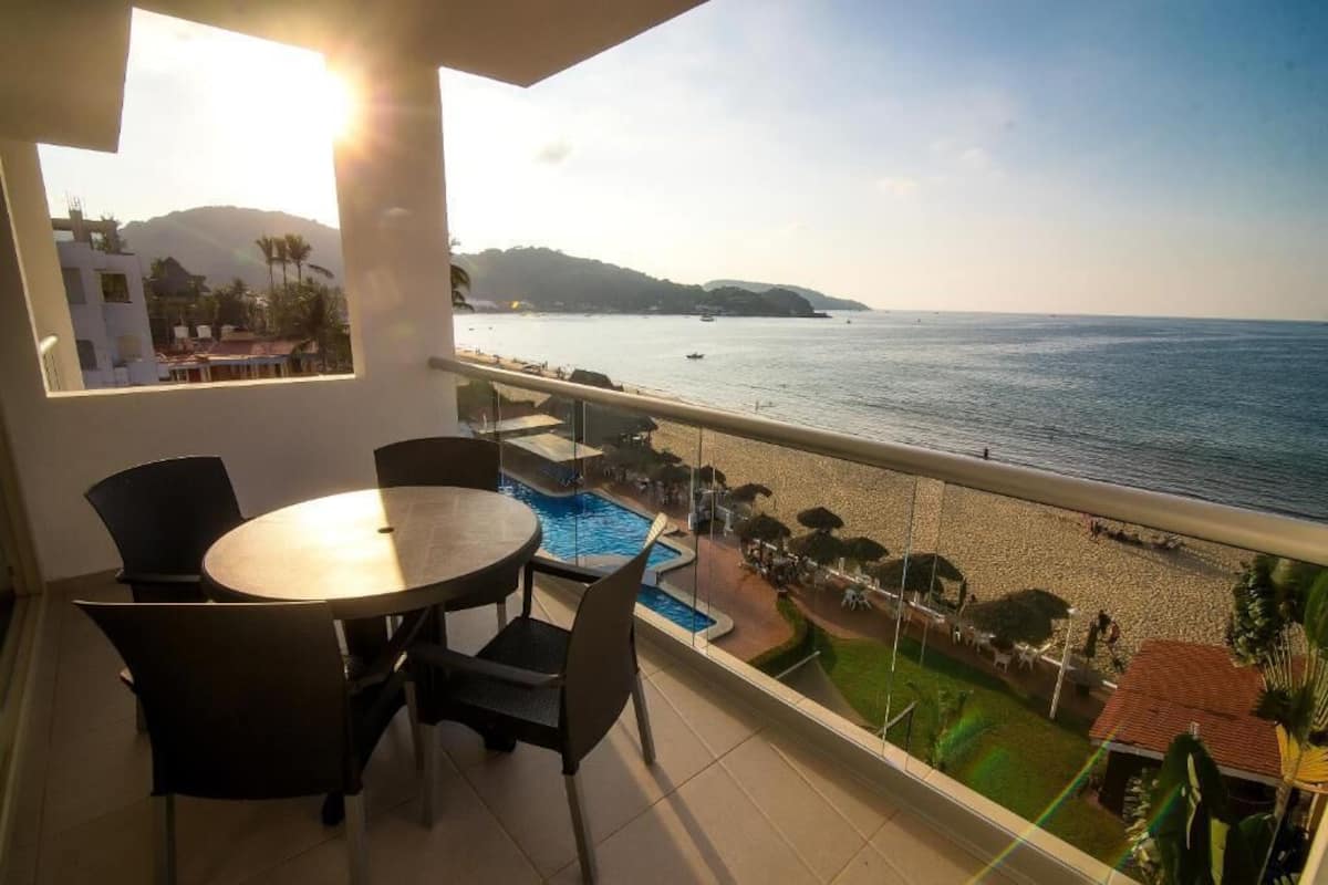 A balcony with a view of the sea, a pool, and a beach area.