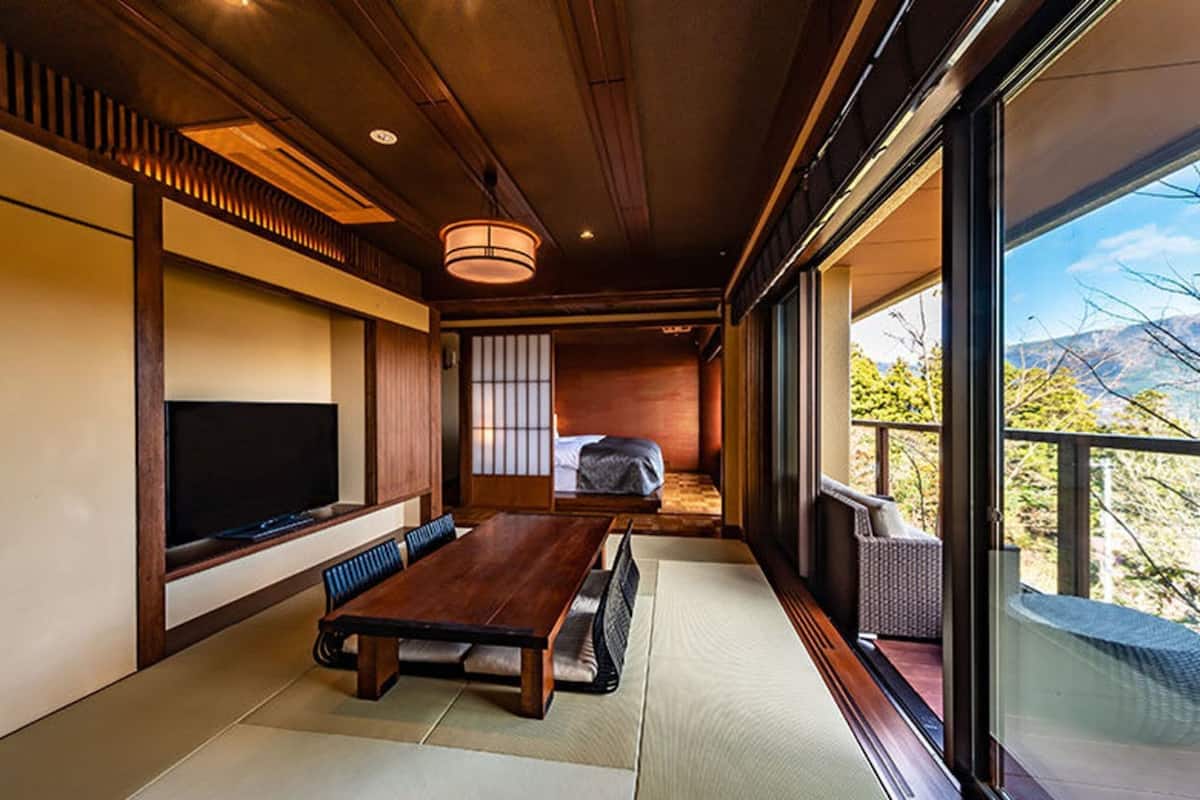 Une pièce de style japonais traditionnel, avec un sol recouvert de tatamis, une basse table en bois, un téléviseur et une porte coulissante donnant sur un balcon avec vue sur la montagne.