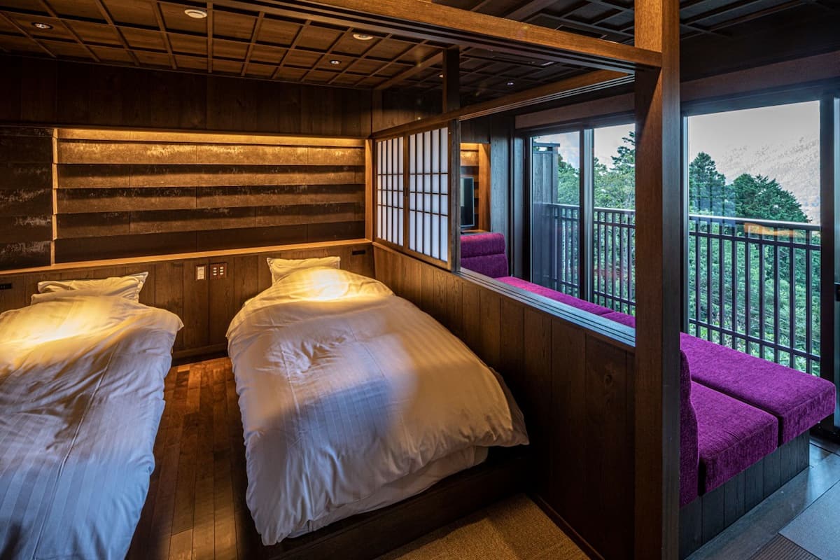 Une chambre avec deux lits, un lambris en bois, un balcon avec des sièges violets et une vue sur la verdure.