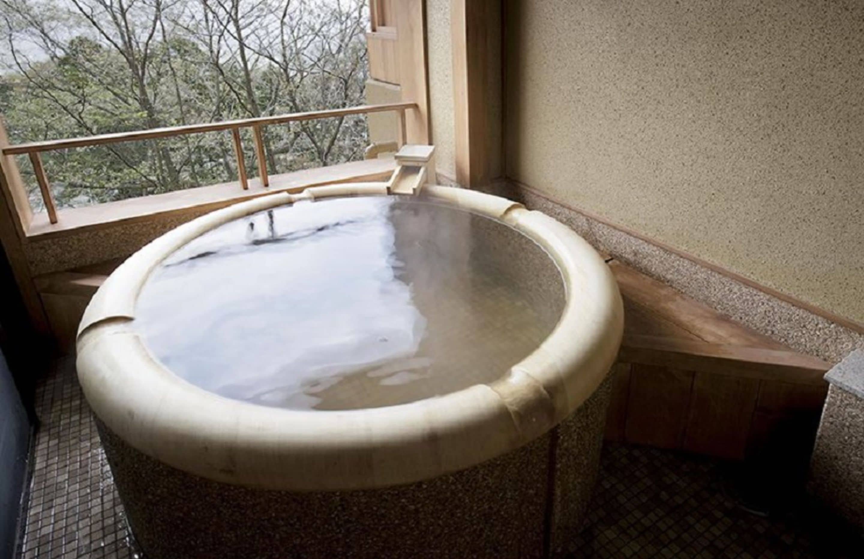 A hot spring bath with a wooden bench and steps leading into it, situated next to a window with a view of trees.
