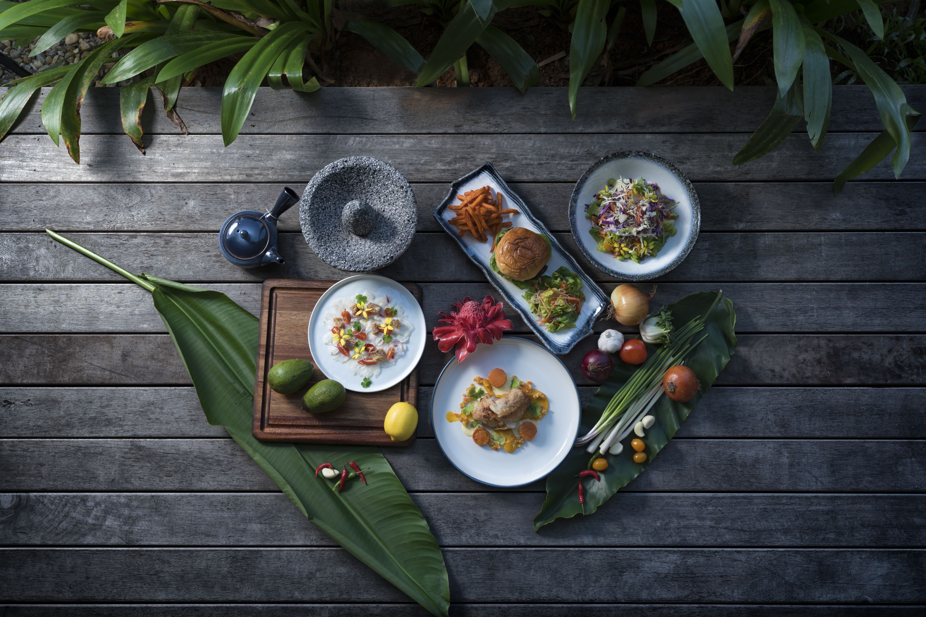 Una mesa de madera con diversos platos, incluyendo una ensalada, una hamburguesa y un plato con pescado, rodeado de verduras y frutas frescas.