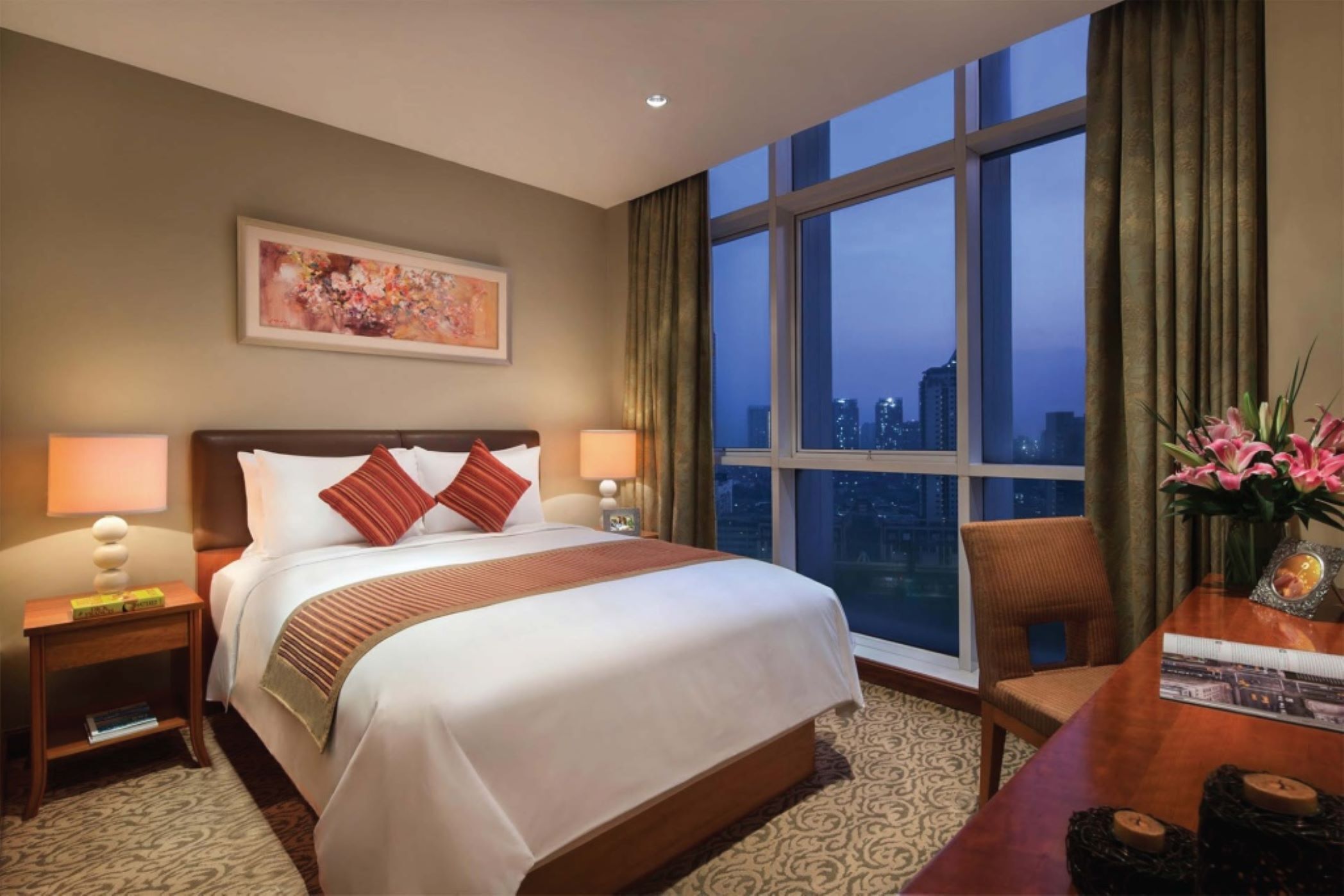A hotel room with a large bed, a desk, a chair, a nightstand, a lamp, a framed picture, and a window with a city view.