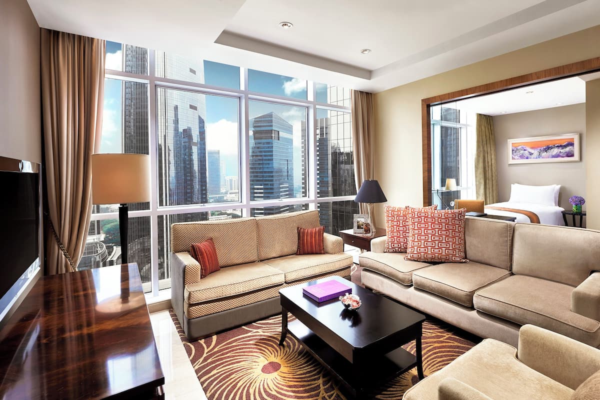 A modern hotel room with a large window offering a city view, a sofa, a coffee table, and a bed with a nightstand.