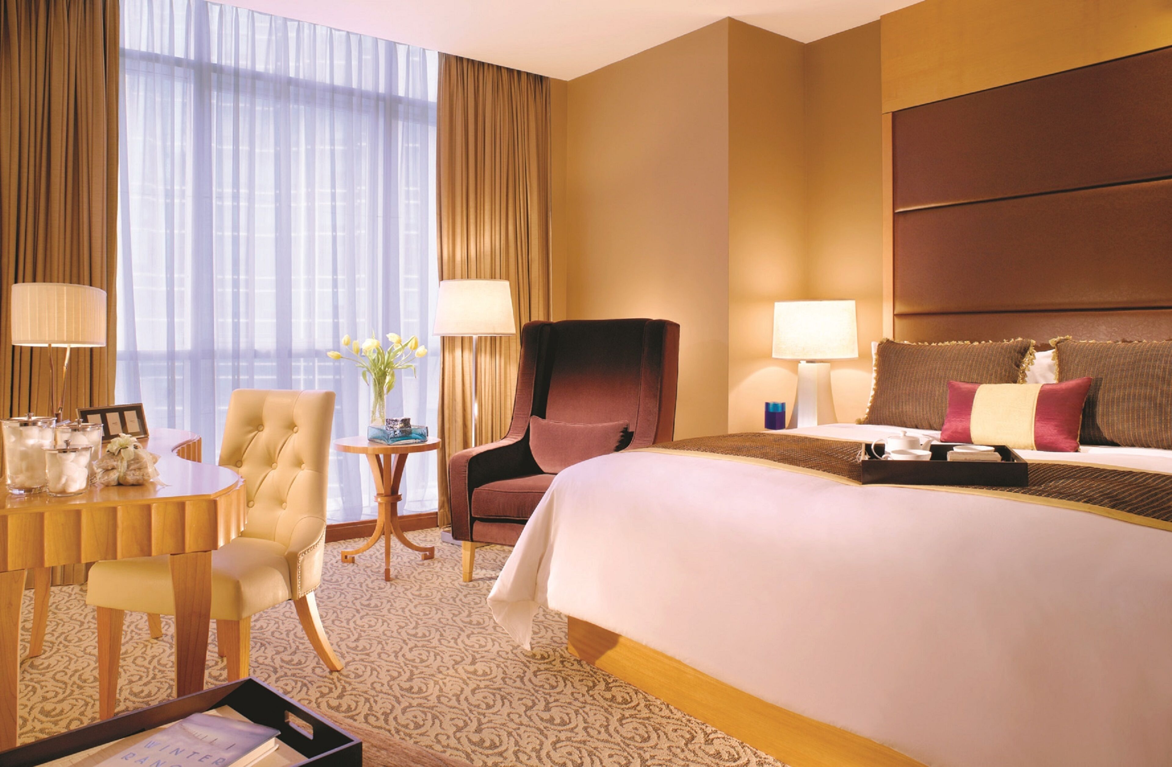 A hotel room with a large bed, a desk with a chair, a small table with a lamp, and a view of the city through large windows.