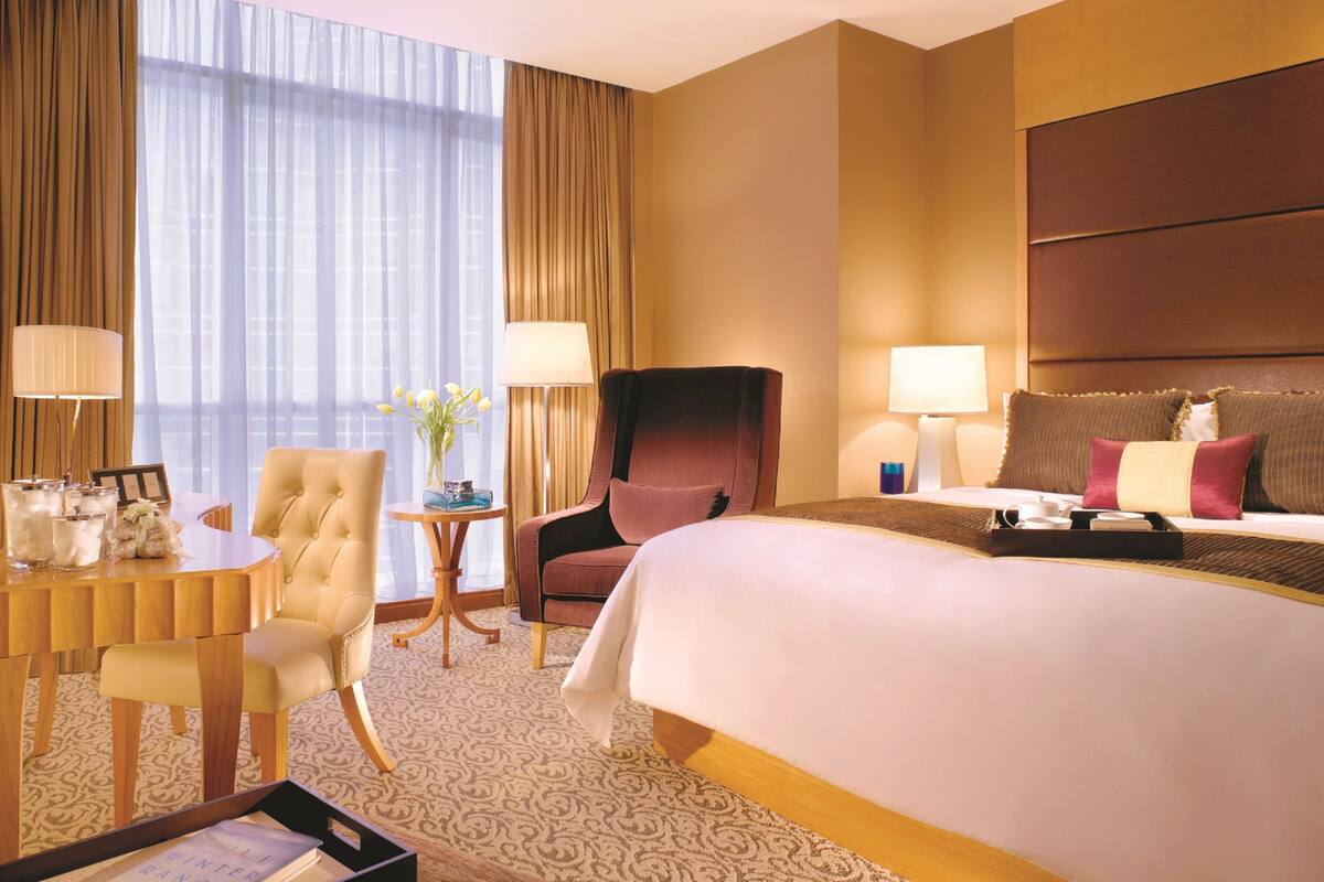 A hotel room with a large bed, a desk with a chair, a small table with a lamp, and a view of the city through large windows.