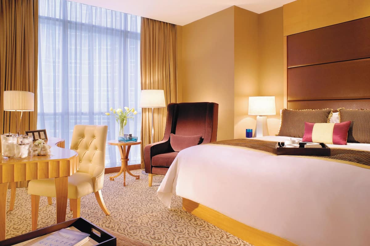 A hotel room with a large bed, two armchairs, a small table with a vase, and a desk with a lamp.